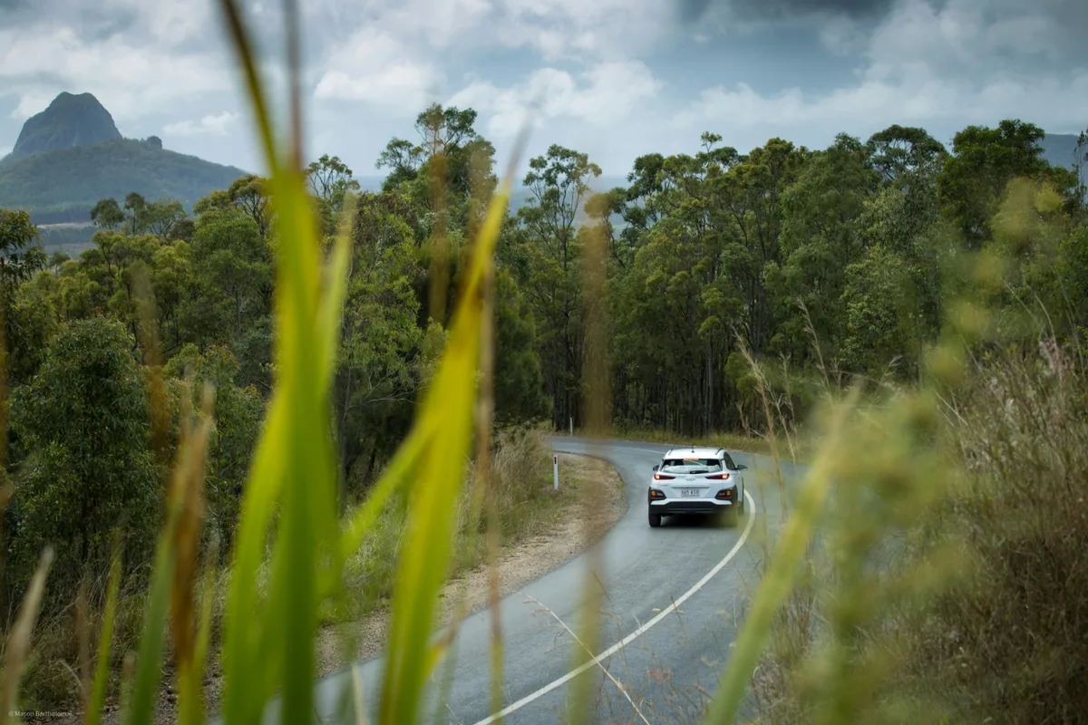 A white car driving on a mountain road seen through tall grass.