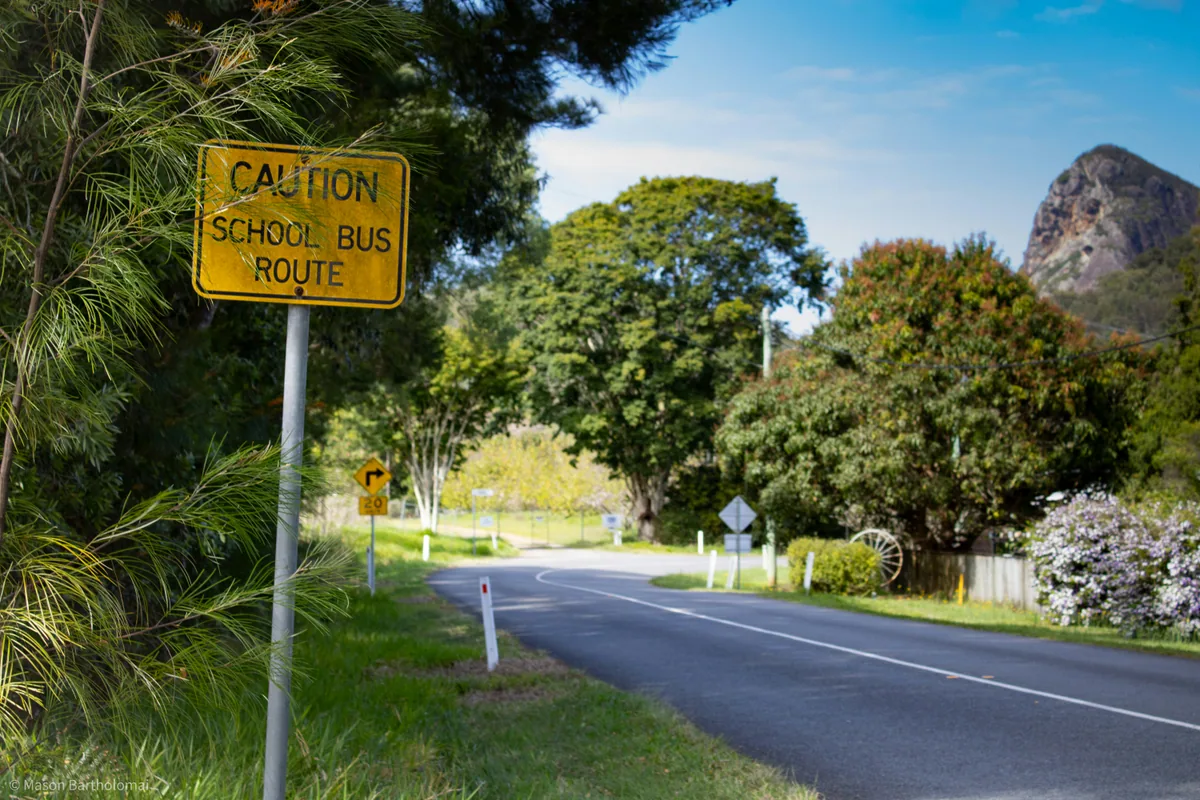 A yellow school bus warning sign beside a quiet road in the Glass House Mountains.